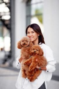 Mujer alegre sostiene dos perros caniches adorables, sonriendo mientras está de pie al aire libre